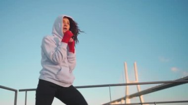 Kapüşonlu gölge boksu giyen atletik kadın boksör. Dişi kickboksör kanca çalışıyor. Elleri kırmızı boks bandına sarılı. Okyanus köprüsü ve mavi gökyüzü arkaplanı ile çalışma 