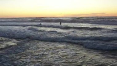 Dalgıç kıyafetli bir grup sörfçü güneş batarken Atlantik Okyanusu siluetinde dalgalara biniyor Carcavelos plajı yaz akşamında. Amatör sörfçüler grubu