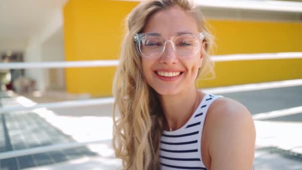 若い美しい金髪の白人女性が眼鏡をかけ スマートフォンを使って外に座って甘い笑顔 良いバイブと感情 モバイルデジタルデバイスを用いたオンライン通信 — ストック動画