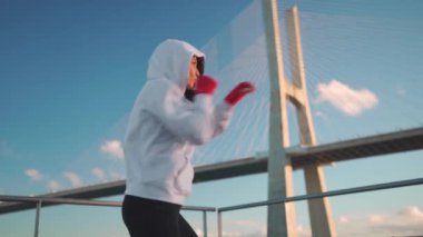 Kapüşonlu kadın boksör, okyanus köprüsü ve mavi gökyüzü geçmişi olan yumruklar eğitiyor. Atletik kadın elleri kırmızı boks sargılarıyla korunuyor. Açık havada Gölge Boksu eğitimi. 