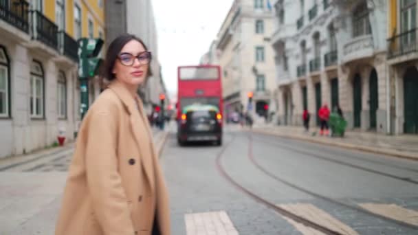 ベージュのコートを着た魅力的な女性とヨーロッパの街を横断する角型メガネは 彼女のスマートフォンを持ち 横に見える 彼女はカメラマンと話していちゃつく 動物園だ 後を追え — ストック動画