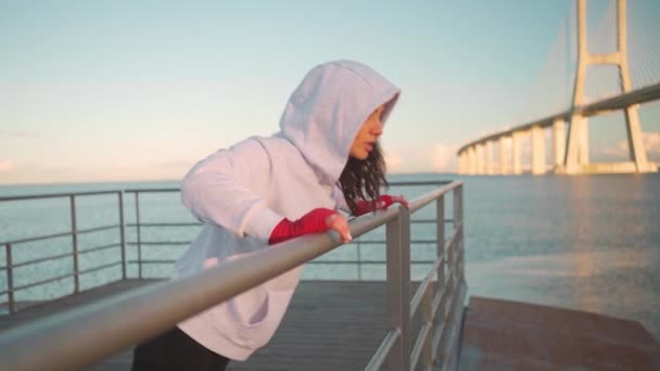 中年女跆拳道手穿着帽衫从有城市海景背景的码头栏杆上做俯卧撑 女运动员的手被红色的拳击带保护着 室外培训 — 图库视频影像