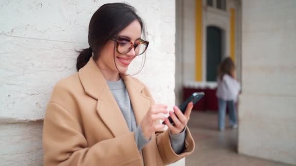 ベージュコートの若い女性と角型メガネは 通りの建物の近くに立っている間 携帯電話でメッセージをチェックします カメラを見ながら携帯電話や笑顔でメッセージを受け取る女性の幸せ — ストック動画