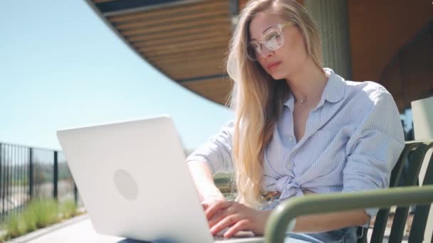 带笔记本电脑的自由人坐在室外咖啡厅的阳台上 戴着眼镜的金发女性穿着休闲装 在户外使用笔记本电脑 使用笔记本电脑从事远程工作的妇女 — 图库视频影像