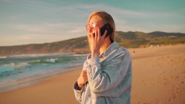 夏休みの間 ビーチに立っているスマートフォンでビデオ通話をする女性観光客 海の近くの携帯電話で話してサングラスとデニムシャツを持つ笑顔の女性 外の技術を使って — ストック動画