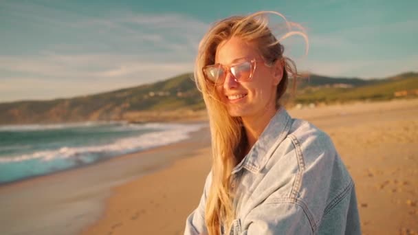 女性は山を背景にビーチに立って海を楽しむ 夕日の海の景色を楽しむサングラスやデニムシャツの女性観光客 夏のビーチ休暇と旅行 — ストック動画