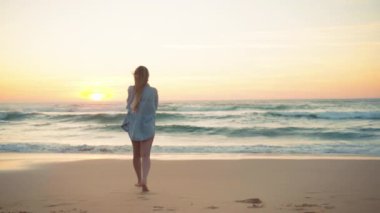 Bikini ve kot gömlekli bir kadın, kumlu sahilde okyanusa doğru yürüyor. Uzun sarı saçlı zayıf kadın gün batımının tadını çıkarıyor. Denizde romantik bir tatil. Arka plan