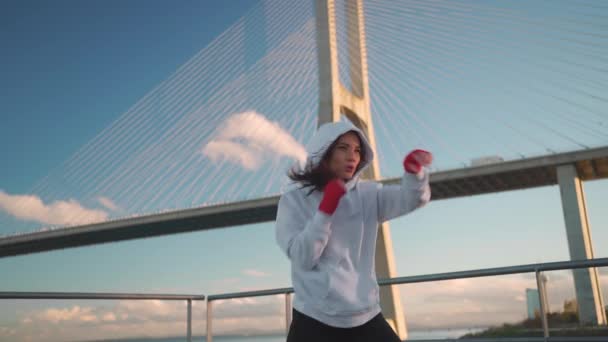 海の橋と青い空の背景を持つ赤いボクシングテープで彼女の手でボクシングのフーディーシャドウボクシングの女性 彼女のジャブとパンチを完璧にする外部の女性アスリート戦闘機トレーニング — ストック動画