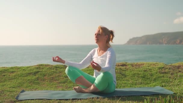 禅修瑜珈在自然界中的修行 一位坐在荷花中沉思的老妇人在海滩上摆姿势 夏日海滩度假期间阳光明媚的早晨沉思 健康积极的生活方式概念 — 图库视频影像