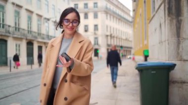 Palto ve çerçeveli gözlüklü genç bir kadın Avrupa sokaklarında yürürken cep telefonundan mesajlar okuyor ve gülümsüyor. Dışarıdaki teknolojiyi kullanarak. Cep telefonu kullanan zarif bir bayan. Çekimi takip et