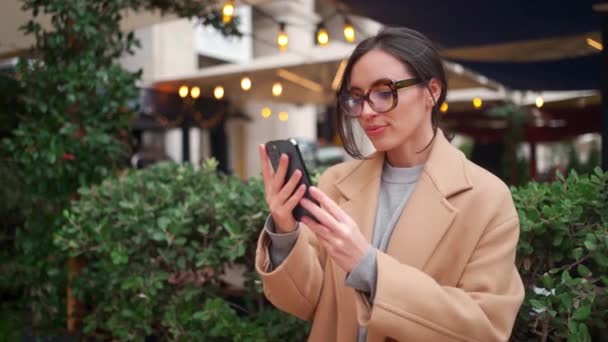 Жінка Пальто Окулярах Читає Повідомлення Мобільному Телефоні Стоячи Біля Декоративного — стокове відео