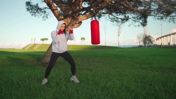赤いボクシングテープに包まれた彼女の手でスポーツキックボクサーの女性のシャドーボクシング 中年女性キックボクサーはフーディーのトレーニングパンチを着用しています 午前中の公園でのトレーニング 全長について — ストック動画