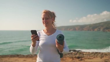 Son sınıf öğrencisi kadın elinde yoga minderiyle okyanus kıyısında akıllı telefonuyla video görüşmesi yapıyor. Sabah yogasından önce cep telefonu kullanan Kafkasyalı mutlu bir kadın. Sağlıklı ve sağlıklı bir yaşam tarzı.