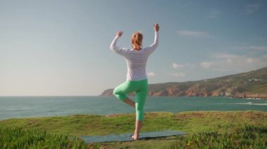 Kıdemli kadın, okyanus kıyısında yoga savaşçısı pozu veriyor. Emekli kadın Vrikshasana pozisyonunda dengede duruyor ve okyanus manzarasının tadını çıkarıyor. Güneşli bir günde doğada spor yapmak. Sağlıklı yaşlı yaşam tarzı