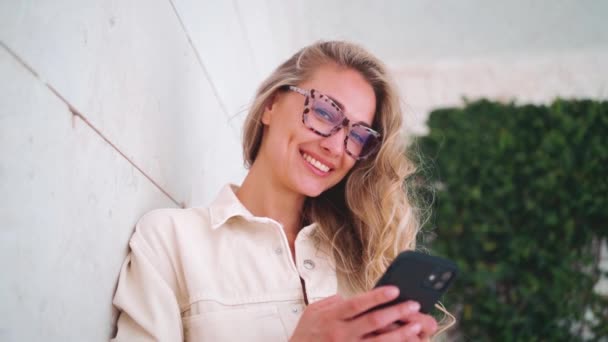 若い魅力的なブロンドの女性が街頭に立って ヒョウのプリントで眼鏡をかけているスマートフォンを保持しています 良い振動とポジティブな感情 携帯電話によるオンラインコミュニケーションの概念 — ストック動画