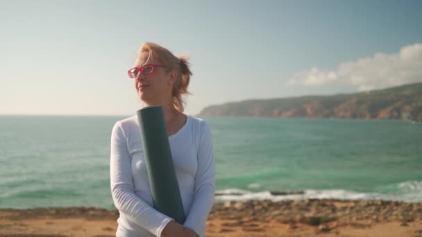 戴着眼镜 拿着瑜伽垫在海滨微笑的快乐的资深高加索女人 退休妇女在晨练瑜伽前享受自然景观 感觉和谐健康健康的老年生活方式概念 — 图库视频影像