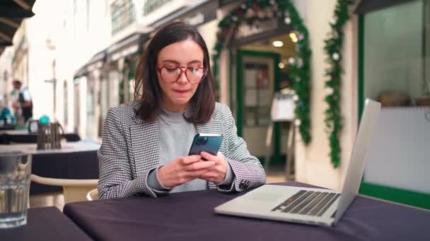 女性博主在她的手机上输入新文章 迷人的年轻女性穿着夹克 戴着眼镜 坐在户外咖啡馆里 在笔记本电脑和智能手机上工作 街头咖啡馆的在线交流 — 图库视频影像