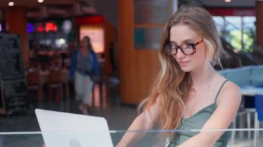 Gözlüklü sarışın kadın uzaktan çalışıyor, bilgisayarla kahve dükkanında tezgahta oturuyor. Laptop ekranına bakarken gülümseyen kız öğrenci