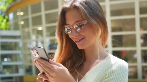 夏の日に公園のカフェの背景に立っている携帯電話を使用している女性 ハッピー 白人女性 メガネ 携帯電話 デバイス ブラウジング インターネット — ストック動画
