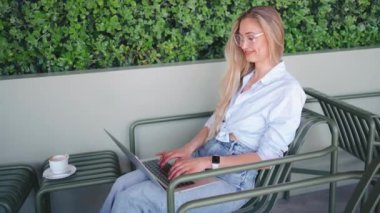 Açık kafe terasında dizüstü bilgisayarı olan serbest çalışan biri. Dışarıda dizüstü bilgisayar kullanan gözlüklü sarışın bir kadın. Orta açı, yüksek açı görüntüsü. Uzak görev için dizüstü bilgisayar kullanan kadın.