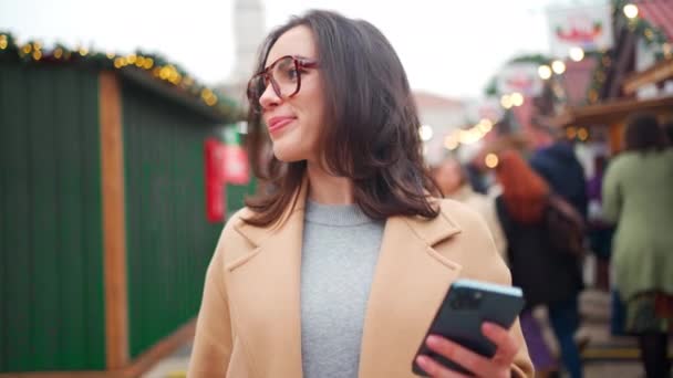在温暖的冬日 穿着米色外套 戴着角边眼镜的优雅的年轻女子在欧洲的圣诞市场上拍照 迷人的女人 手里拿着手机 看着相机 面带微笑 — 图库视频影像