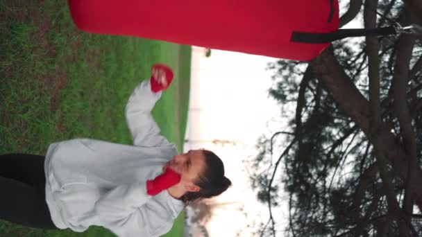 在公园里练拳击时 戴着帽衫的跆拳道女子在打沙袋 田径女拳击手的手被红色的拳击带包裹在室外训练 女性健身动机 垂直录像 — 图库视频影像