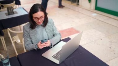 Ceketli ve gözlüklü bir iş kadını açık kafede oturuyor ve dizüstü bilgisayar ve akıllı telefon üzerinde çalışıyor. Cep telefonuna mesaj yazan bir kadın. Sokak kafesinde internet bağlantısı. Yüksek açı