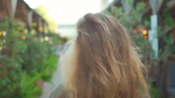 若い白人ブロンドの少女がカフェの屋外テラスを歩き 笑顔でカメラに向かう姿を背景にしています 夏の夕方に緑とストリートガーランドの背景にメガネをかけた幸せな若い女性 — ストック動画