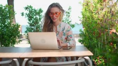 Çekici iş kadını güneşli yaz gününde terasta dizüstü bilgisayarıyla oturuyor. Gözlüklü, güzel elbiseli, esintiden hoşlanan modern kadın serbest yazar. Hoş bir atmosferde uzak çalışma