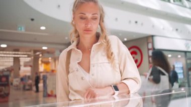 Modern alışveriş merkezinde cam vitrin mücevheri ve aksesuar arayan zarif orta yaşlı beyaz kadın. Mağazada mücevher seçen beyaz sarışın kadın. Pencere gösterimleri ve alışveriş arkaplanı