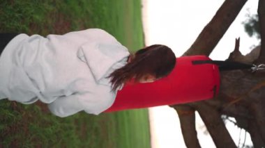 Kapüşonlu kadın parkta boks yaparken kum torbasına vuruyor. Atletik kadın boksör elleri açık havada antrenman yaparken kırmızı boks kasetlerine sarılmış. Kadın fitness motivasyonu. Dikey video