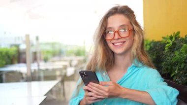 Gözlüklü genç, güzel bir sarışın bir yaz günü bir kafede oturmuş elinde akıllı bir telefonla kameraya hoş bir şekilde gülümsüyor. İyi hisler ve hisler. Bir mobil dijital aygıt kullanarak çevrimiçi iletişim 