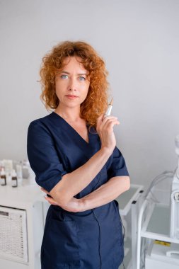 Tıp bürosundaki kadın kozmetik uzmanı. Önlük giyen kızıl saçlı kadın dermatolog hastanede bir ampul tutuyor. Cilt bakımı uygulayıcısı lacivert tıp üniformalı estetik klinik ofisinde duruyor.