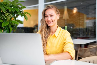 Freelance woman using laptop outdoor. Freelance worker sitting at cafe terrace remote working. Professional freelancer wearing yellow jacket sitting under green plant at cafe table with computer.  clipart