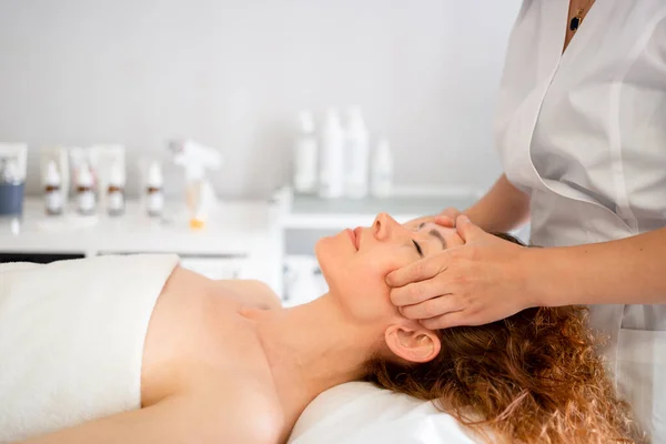 Women takes aesthetic procedure, covered white towel lays down in cosmetology clinic. Patient of Beaty clinic having face massage. Female cosmetologist gives aesthetic treatment to female client.
