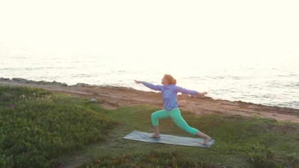 退休妇女日落时在海滩上做瑜伽 一个成熟的女人在海滨练习瑜伽 身体护理 身体活动的概念 空中景观 — 图库视频影像