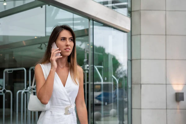 Business woman has phone call outdoors. Portrait of successful female talking on cellphone outside of bank. Professional business lady wearing white dress has important phone conversation.