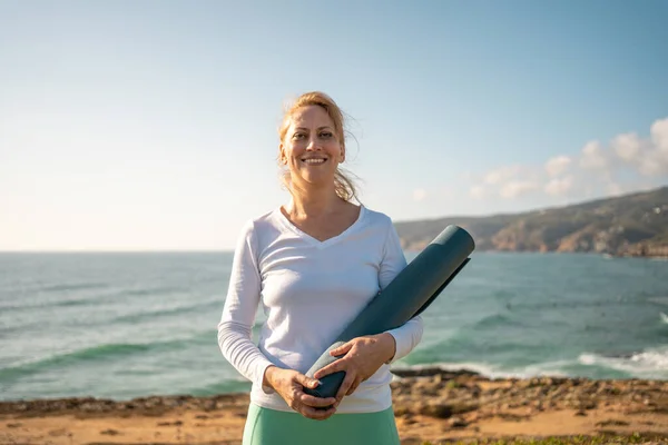 Senior female holding fitness mat on beach. Pensioner woman smiling while holding yoga mat with both arms, coast line on backgrounds. Mature sporty person standing on ocean shore ready for yoga