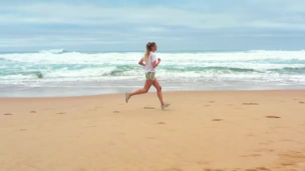 空中撮影された若い女性は 夏の朝に美しいビーチでジョギングします 海岸を走る屋外で運動するブロンドの白人女性 健康的なライフスタイルとスポーツのコンセプト — ストック動画