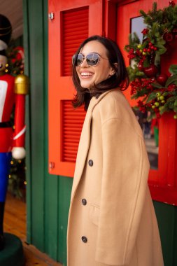 Avrupa şehrinde Noel tatili günü yürüyüş yapan Kafkasyalı mutlu kız. Esmer kadın şık bir ceket, ceket ve güneş gözlüğü takıyor. Olumlu duygular yaşam tarzı kavramı