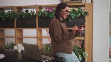 Modern çalışma sahasında kadın girişimci. Kahve molası sırasında gözlük takan iş kadını cep telefonundaki mobil uygulamaları kontrol ediyor. Çalışma odasında dizüstü bilgisayar ve cep telefonu kullanan bir kadın 