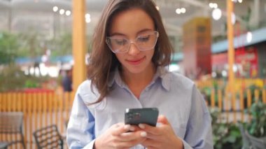 İş kadını cep telefonuyla yazı yazıyor ve gülümsüyor. Alışveriş merkezinde gözlüklü bir iş kadını geziyor ve mobil uygulama ile mesaj yolluyor. Modern yaşam tarzı kavramı