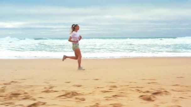 航空追跡ショット 海岸線に沿ってジョギングブロンドの女性 海岸を走るアウトドアで運動する若い白人女性 健康的なランニングとアウトドアの運動の概念 サマーアクティブ — ストック動画