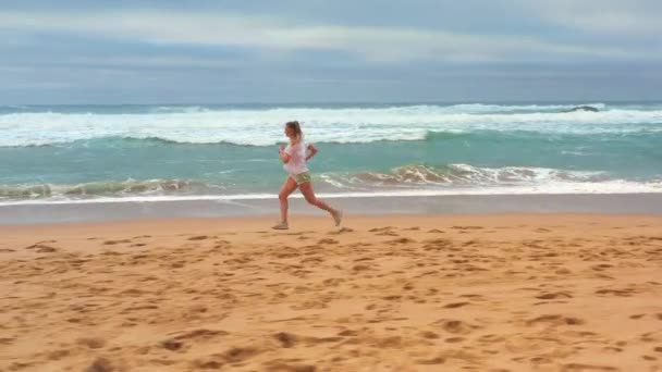 午前中に海岸線に沿ってジョギングする若い女性を航空追跡 ブロンドの白人女性は屋外で運動します 健康的なランニングとアウトドアの運動の概念 サマーアクティブ — ストック動画
