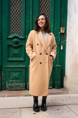 Arkadaş canlısı, güler yüzlü çekici kadın Avrupa şehrinin arka planındaki yeşil çit üzerinde binayı terk ediyor. Gözlüklü, şık trençkotlu mutlu bir kadın kameraya bakıp gülümsüyor.