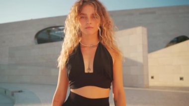 Kıvırcık saçlı bir kadın yazın dışarıda poz veriyor. Siyah elbiseli, rahat sarışın kadın güneş ışınlarının altında kameraya bakıyor.