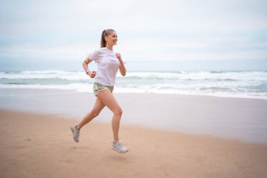 Okyanus kıyısında koşan bir kadın. Beyaz tişörtlü ve yeşil şortlu doğa kadını deniz kıyısı boyunca koşuyor. Atletik kadın sabahları kumsalda koşuyor.