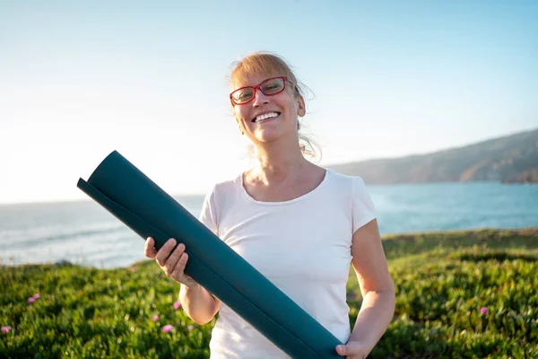 Happy elderly woman wearing glasses stand with yoga mat smiles on ocean shore. Retired woman enjoys views of nature before her morning yoga on beach. Concept of healthy lifestyle for elderly person