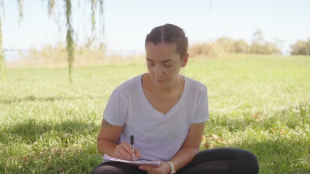 有笔迹的女人坐着写莲花的姿势 在笔记本上写着字 在草地上 在野外 在田野里 夏天的日子 女作家穿着白衬衫 写着便条 — 图库视频影像