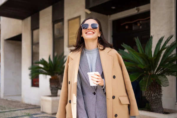 Happy woman in sunglasses and coat walks out of hotel smiling. Young adult woman walks looking up in sunglasses dressed in coat and smiling autumn. Female with cup of coffee smiles at autumn outdoors